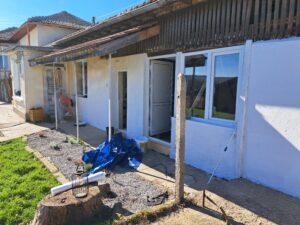 A person is painting the exterior wall of a white guest house. Supplies, including a blue tarpaulin, paint tins, and poles, are scattered nearby. The house at Muddy Puddles Farm has a tiled roof and windows, brightly lit by the sunlight.
