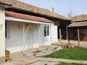 A small white guest house with a red-tiled roof, glass door, and a porch with a wind chime. Set on a green lawn in Kovachevets, it features a tree stump, stacked firewood, and a shed in the background.