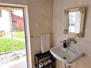 A small tiled bathroom at Muddy Puddles Farm's guest house in Kovachevets, featuring a white basin, ornate mirror, towel rail with a white towel, and black shelf with essentials, with a door open to a sunny grassy garden.