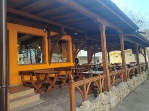 A wooden outdoor terrace with empty tables and benches sits under a wooden roof, attached to the bright orange walls and large windows of the Muddy Puddles Farm guest house. The area appears calm and inviting.