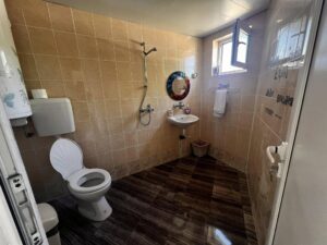 A bathroom in the Muddy Puddles Farm guest house features beige tiled walls, a toilet, a wall-mounted washbasin with mirror, showerhead, towel rail with white towel, window, and a brown waste bin on the dark tiled floor.