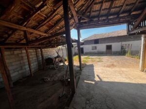 View from inside a rustic, open wooden shed at Muddy Puddles Farm towards a sunny courtyard with old buildings. A metal wheelbarrow rests in the shade; uneven ground blends dirt and concrete. Shadows fall gently across this charming farm stay scene.