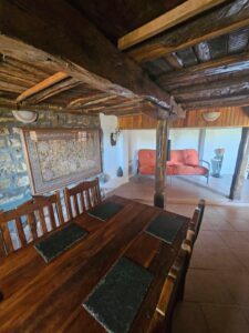 A rustic dining room at Muddy Puddles Farm features a wooden table and chairs, stone walls, and exposed beams. In the background, an orange sofa, standard lamp, and decorative tapestry add cosy farm stay charm to this guest house setting.