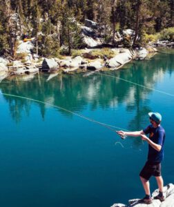 A person in a blue shirt, shorts, and cap stands on a rock near Kovachevets, casting a fishing line into a clear, blue lake surrounded by rocks, trees, and forested mountains—a perfect retreat after a day at the nearby farm stay.