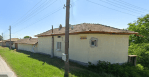 A single-storey yellow guest house with a tiled roof sits beside a grassy roadside in Kovachevets. Power lines and a utility pole run alongside, bushes grow near the building, and there is a small sign on the wall.