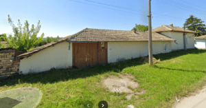 A long, single-storey white guest house with a brown tiled roof and wooden double doors sits beside a grassy roadside under a clear sky, with a telephone pole and trees in the background—perfect for your Muddy Puddles Farm stay.