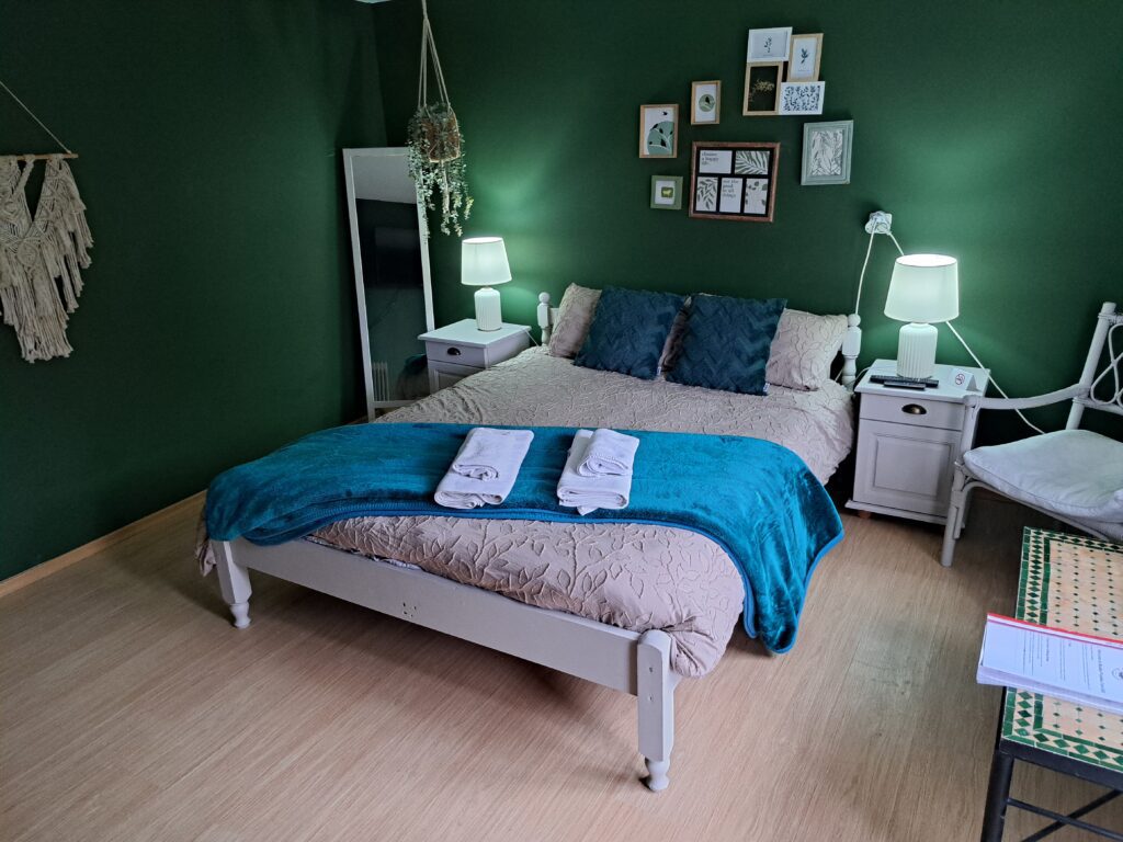 A cosy bedroom at Muddy Puddles Farm in Kovachevets, with dark green walls, a white bed with teal and beige bedding, folded towels, bedside tables with lamps, charming wall art above the bed, a chair, and a mirror in the corner.