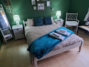 A cosy guest house bedroom in Kovachevets with green walls, a double bed with a beige patterned cover, teal throw and pillows, two white bedside tables with lamps, a chair, framed wall art, and neatly folded towels on the bed.