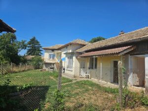 Two old, beige houses with tiled roofs stand side by side at Muddy Puddles Farm in Kovachevets, surrounded by a grassy garden and wire fence, under a clear blue sky with trees in the background.
