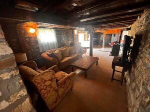 A cosy rustic living room at Kovachevets Guest House features stone walls, a wooden beamed ceiling, a window with sheer curtains, a patterned armchair, soft cushions on the sofa, and a wooden coffee table on a tiled floor.