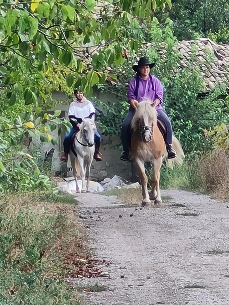 Two people ride horses down a dirt path at Muddy Puddles Farm in Kovachevets, surrounded by greenery. The rider in front wears a purple shirt and black hat; the other wears white. Trees and an old, tiled-roofed building appear in the background.