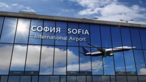 The mirrored glass exterior of Sofia International Airport reflects a blue sky as a commercial aeroplane flies past the building, welcoming travellers from destinations like Muddy Puddles Farm, a popular farm stay near Kovachevets. The airport’s name is in Cyrillic and Latin.