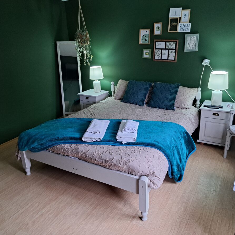 A cosy farm stay bedroom in Kovachevets with green walls, a double bed dressed in blue and cream bedding, two folded towels at the foot, bedside tables with lamps, framed art on the wall, a mirror, and a white chair with a cushion.