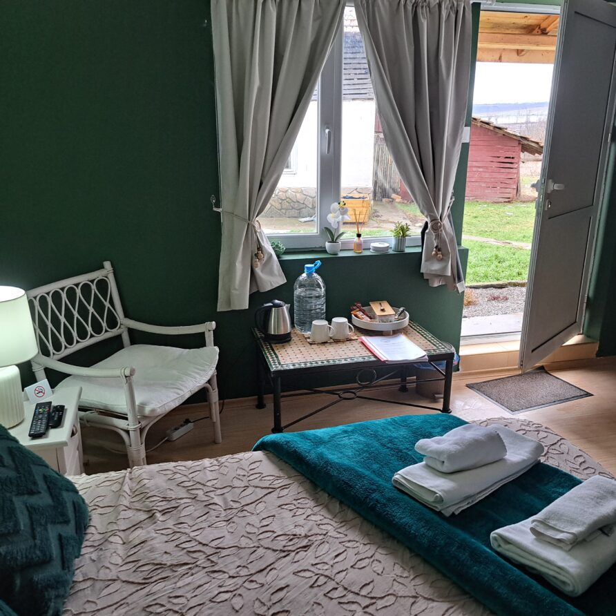 Cosy guest room at Muddy Puddles Farm with a bed, towels, a small table with refreshments, chair, and bedside lamp. A window with light curtains and an open door reveal a grassy outdoor area and wooden shed.