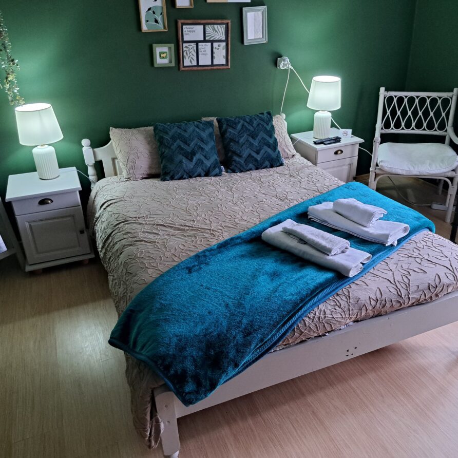 A cosy guest house bedroom in Kovachevets with green walls, a double bed with patterned bedding, dark blue cushions, and a teal throw. Two bedside tables with lamps, a chair, mirror, and wall art complete the decor; towels are folded on the bed.